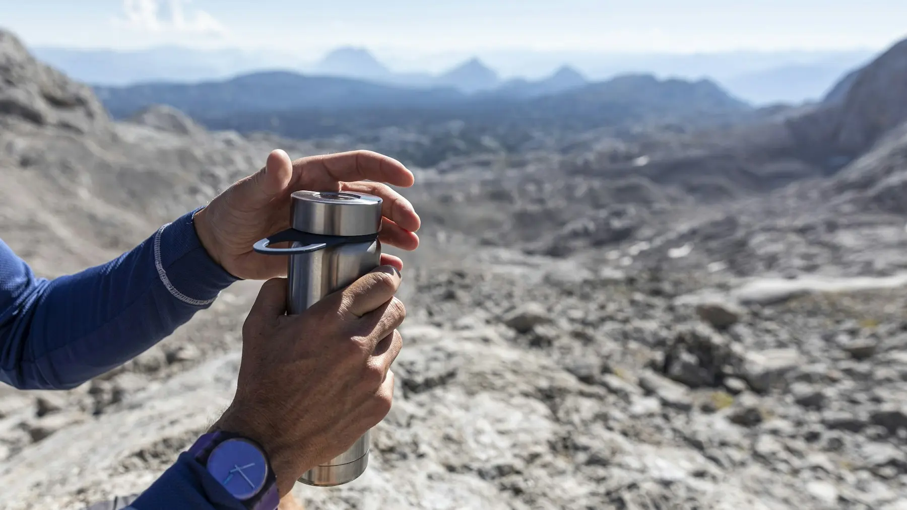 A hiker holding a reusable metal bottle above a mountain landscape