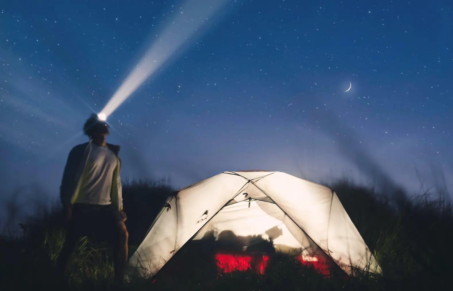 A camper wearing a headlamp beside an illuminated tent under a starry sky